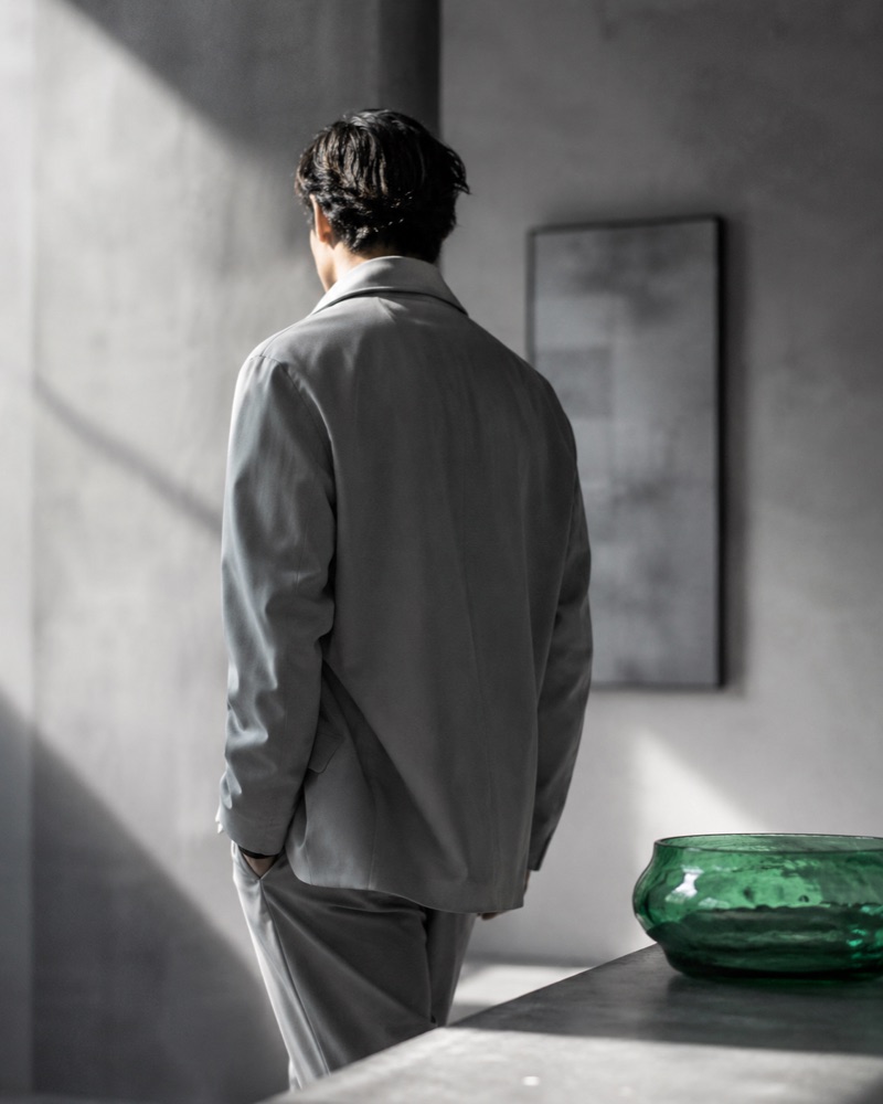 A man in a tailored grey suit in a quiet concrete space, beside a green glass vessel